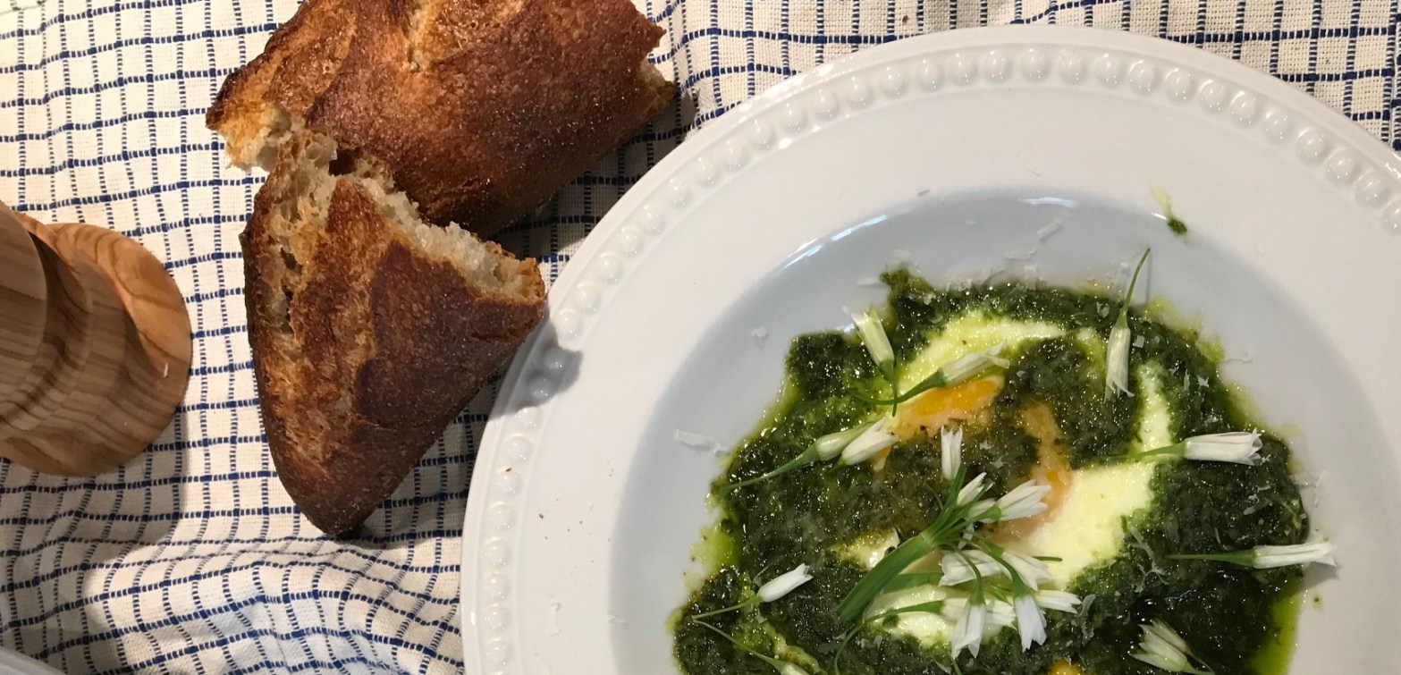 A plate of eggs baked in a green mixture of ramps and olive oil with two pieces of baguette and a wooden pepper grinder on a blue and white checkered napkin